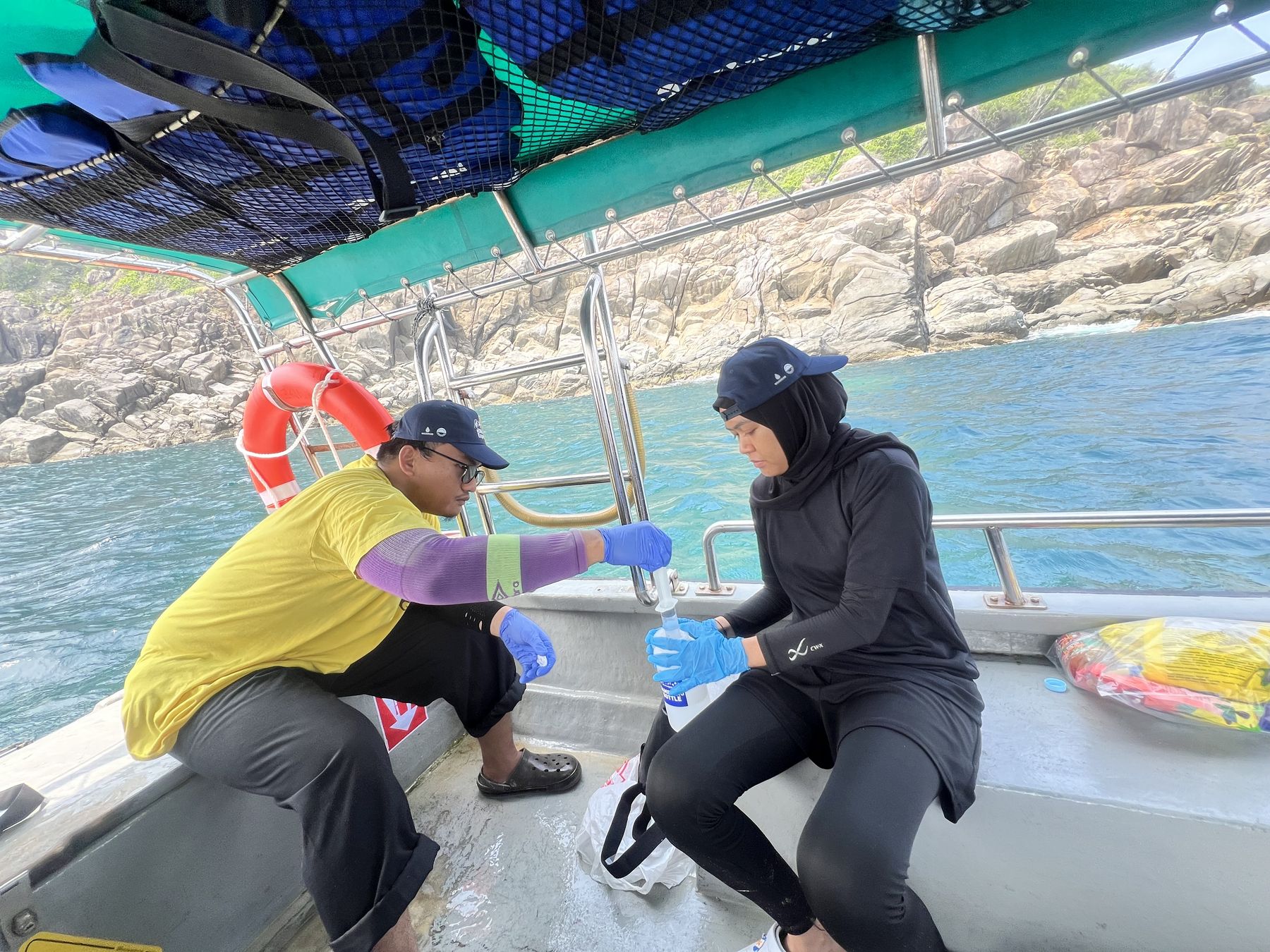 Under intense heat and a scorching sun, two participants fill a sampling syringe from a sampling bottle, preparing for eDNA filtering. Terengganu, Malaysia, September 2026.