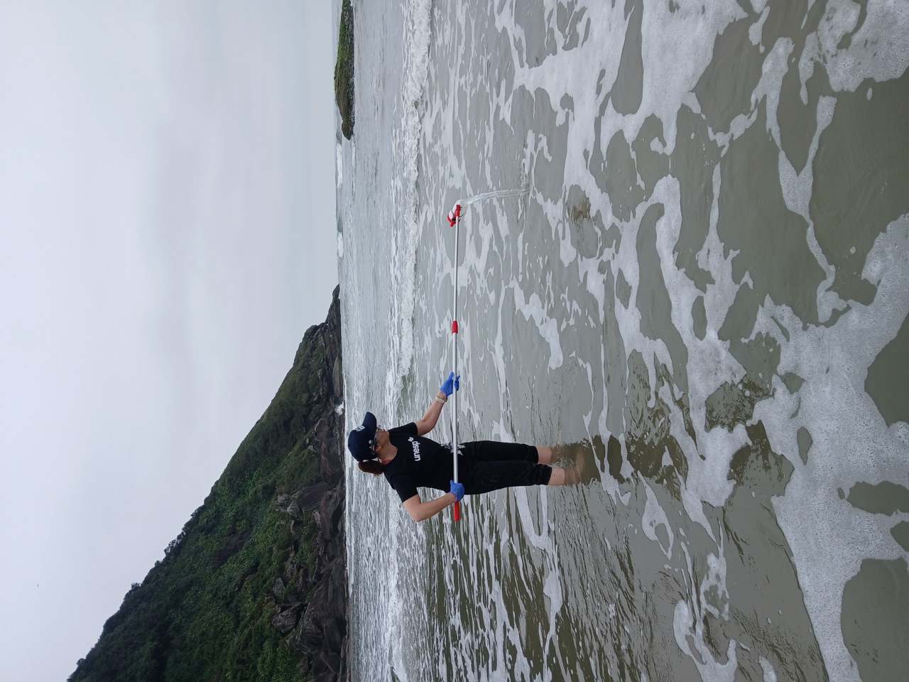 Prof. Giovana Bertini collecting a water sample in the surf at Cananéia–Iguape–Peruíbe, Brazil, August 2026.