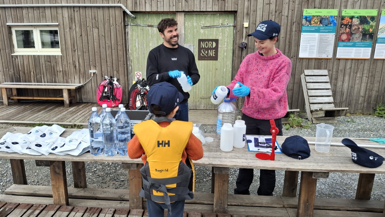 Researchers Amy Mackintosh and Loïc Sanchez and a young helper, preparing bottles and equipment ahead of their first sampling event, in front of the Nord&Ne diving club on the Eastern side of the maelstrom, in August 2026.